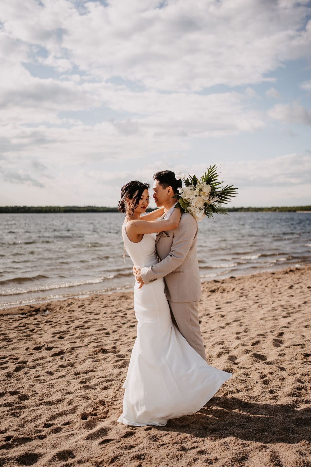 A couple embraces on a sandy beach during their wedding photoshoot with clouds and water in the background.