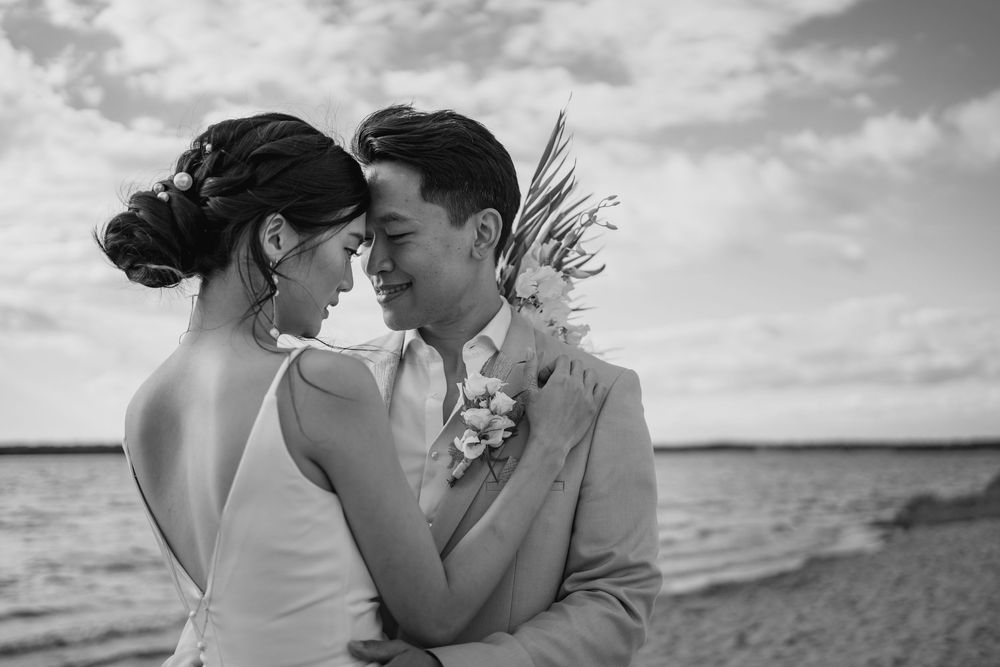 A romantic black and white beachside wedding photo showing a tender moment between newlyweds against an ocean backdrop.