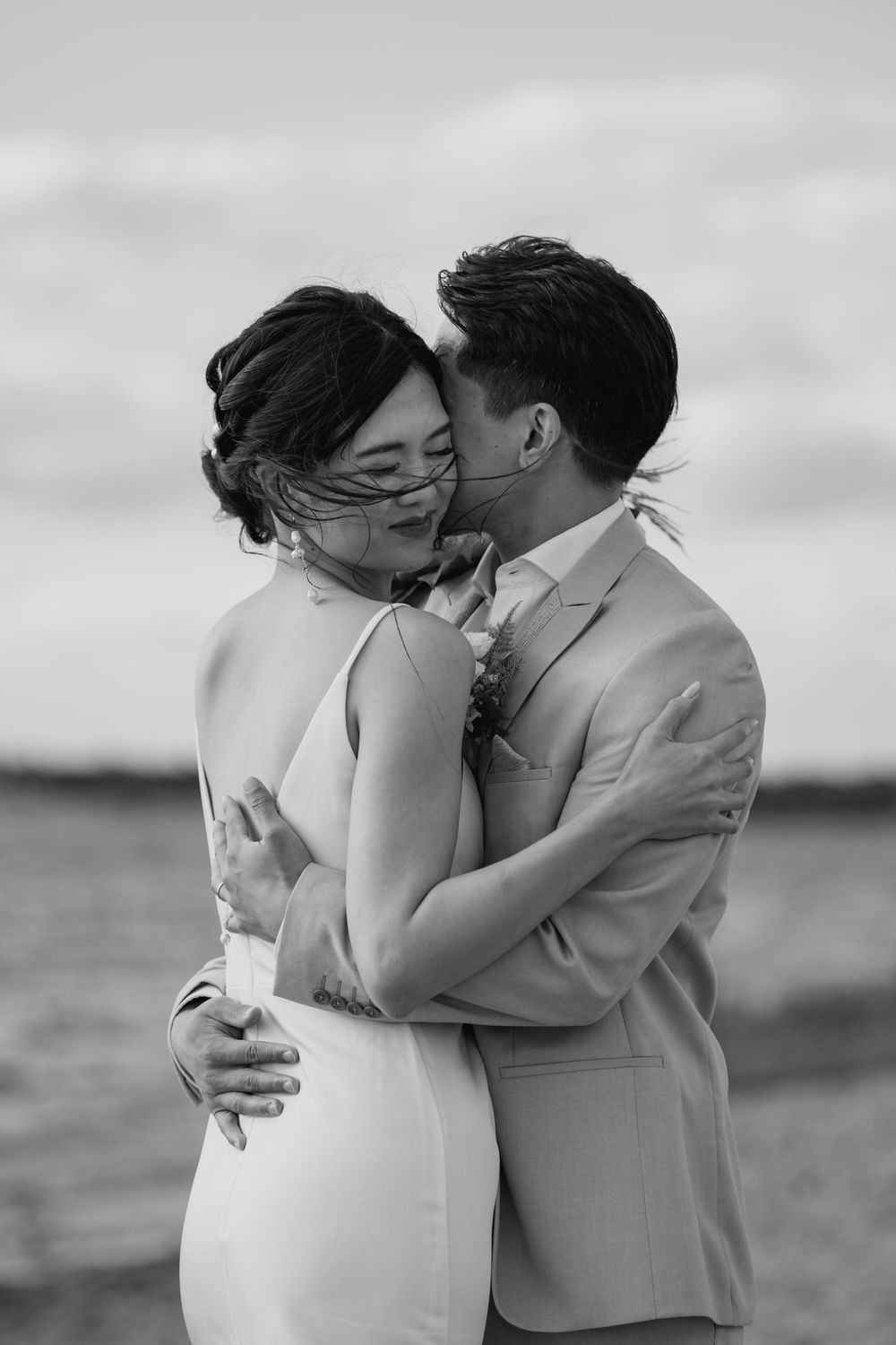 Black and white wedding photo of a tender moment between a couple embracing on a beach with water in the background.
