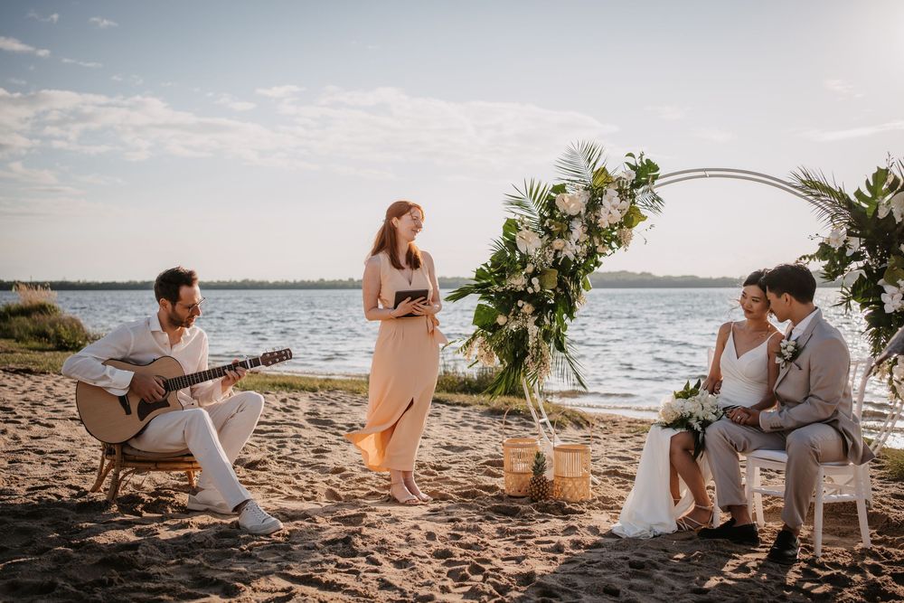 Intimate beach wedding ceremony with acoustic guitar music under a floral arch at sunset by the water.