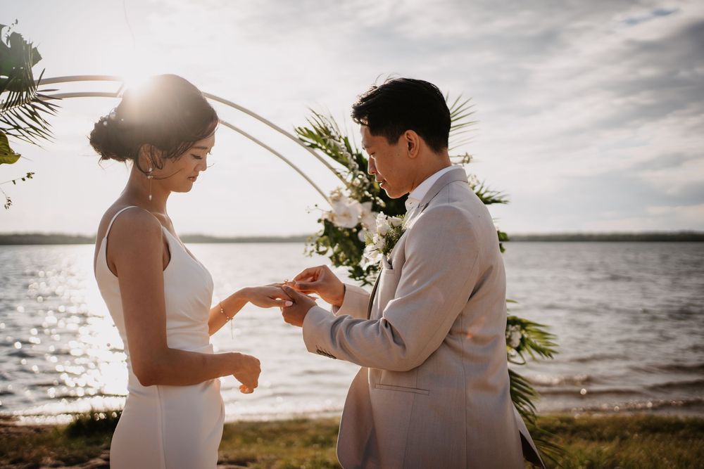 Couple exchanging rings during intimate beach wedding ceremony with ocean waves and cloudy sky in the background.