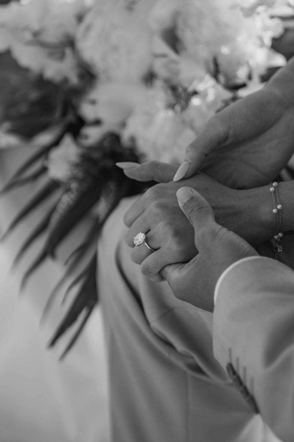Black and white closeup of clasped hands showing engagement ring with floral bouquet in soft focus background.