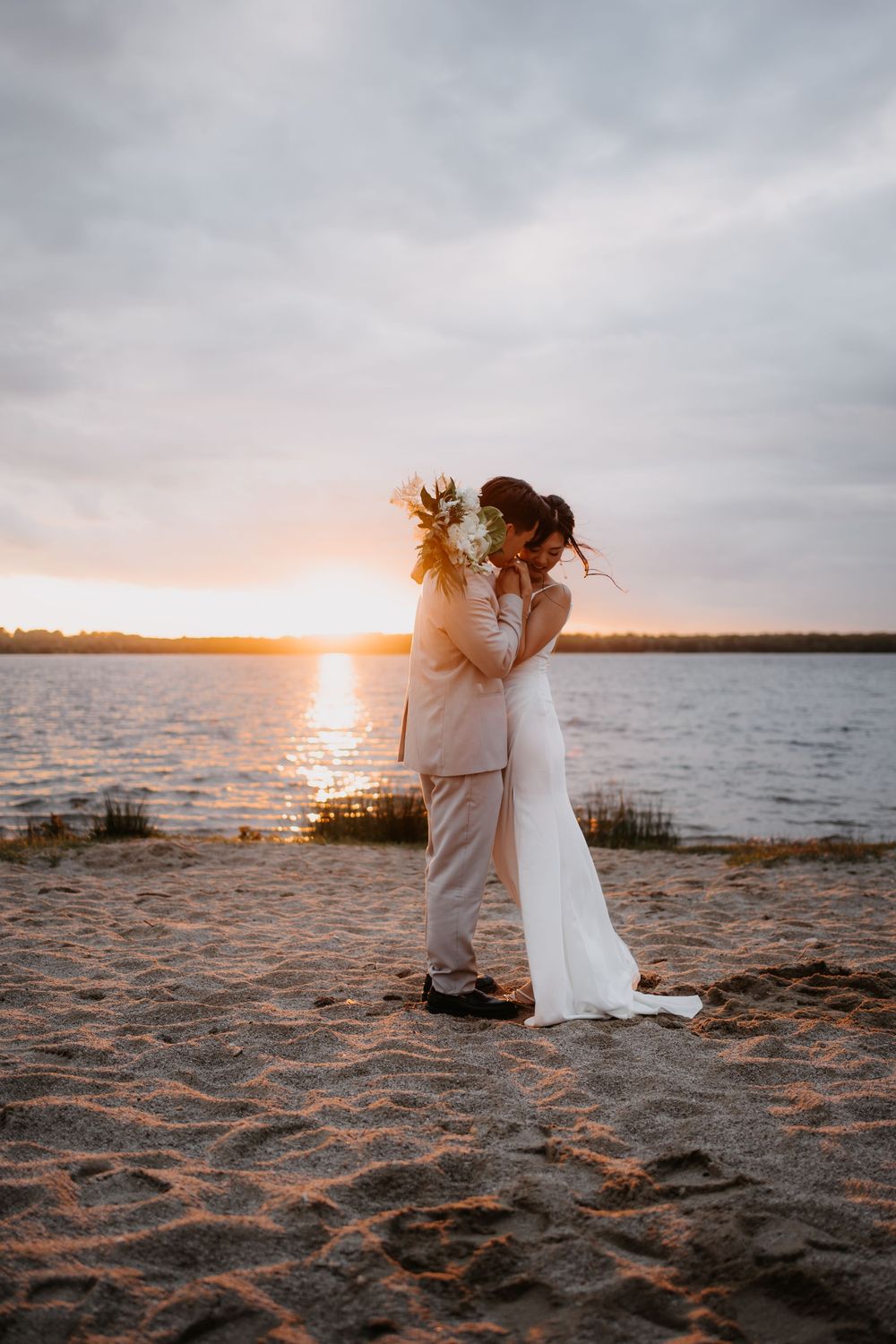 A couple embraces on a sandy beach at sunset, silhouetted against a golden sky and tranquil lake.