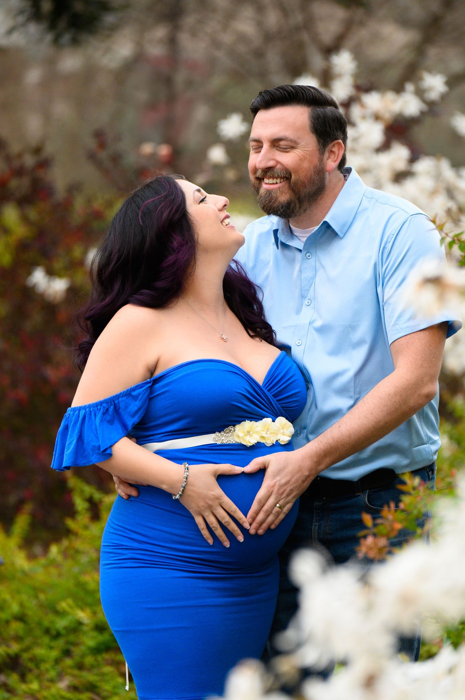 A couple shares a tender moment outdoors during a maternity photoshoot with white blossoms in the background.