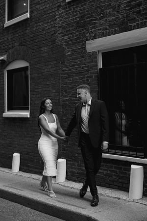 A couple dressed elegantly in wedding attire walk together on a sidewalk by a brick building in black and white.