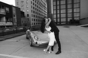 A stylish black and white photo shows a romantic dipping pose next to a vintage Porsche car in an urban setting.