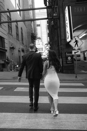 A black and white photo of a couple walking across a city crosswalk dressed in formal attire.
