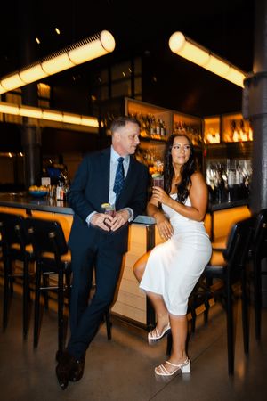Elegantly dressed individuals sharing drinks at an upscale bar with modern lighting fixtures and dark ambiance.