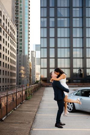 Romantic couple embraces in urban parking garage with downtown buildings and glass architecture in background.