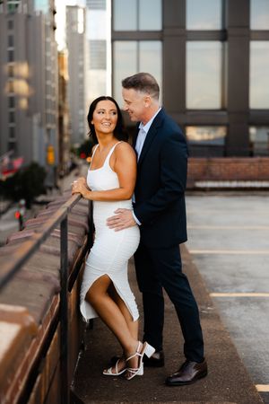 Romantic couple embraces on urban rooftop at sunset, with her in white dress and him in navy suit.