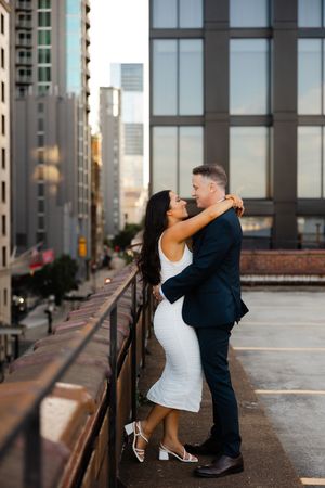 A couple embraces on a rooftop parking garage with the city skyline visible in the background.