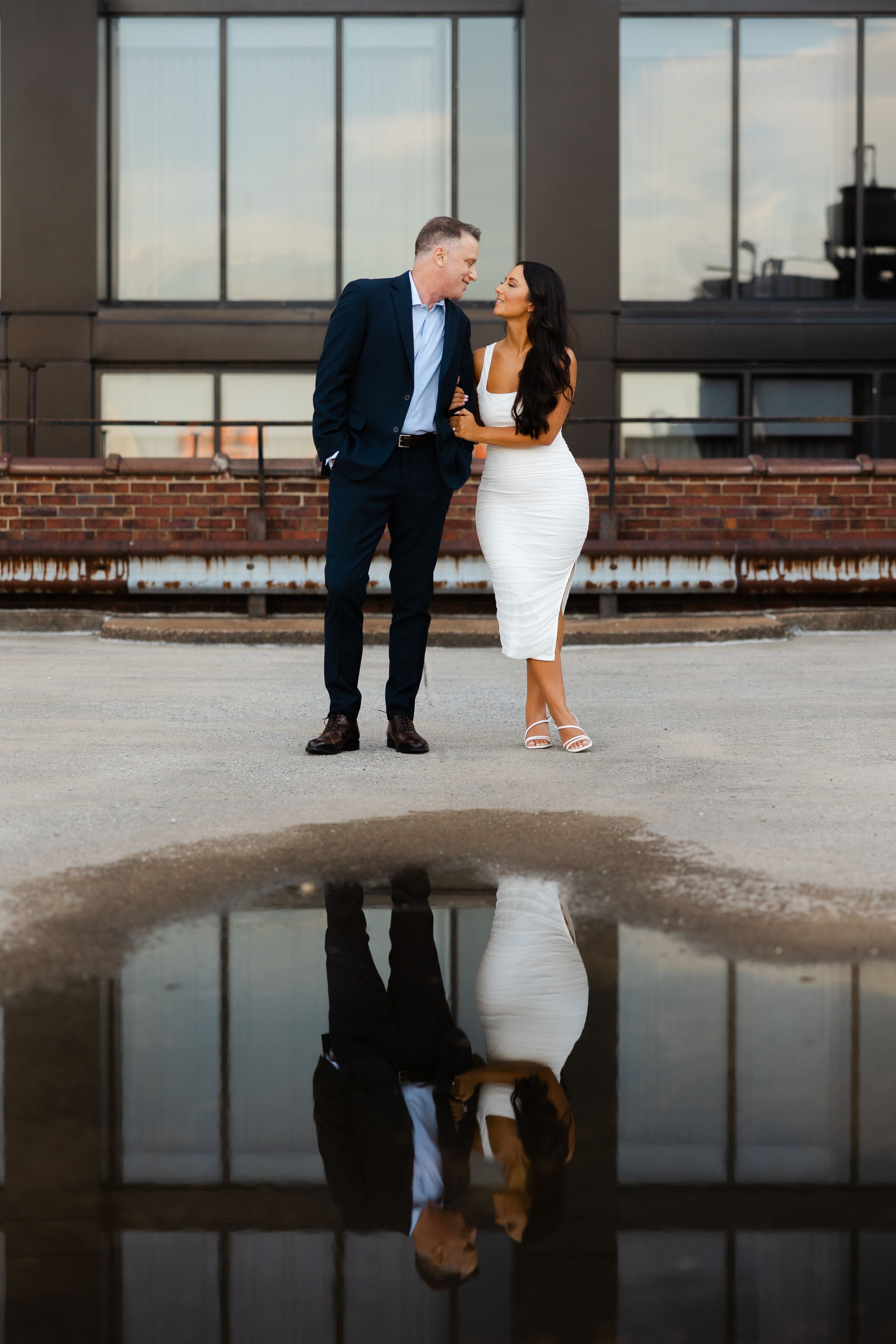 A couple in formal wear reflected in a puddle against an urban architectural setting.