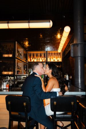 A romantic moment between a couple at an elegant bar with soft lighting and bottles displayed on illuminated shelves.