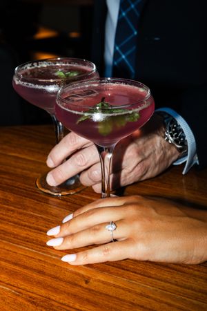 Two purple cocktails garnished with herbs on a wooden bar table with hands holding elegant martini glasses.