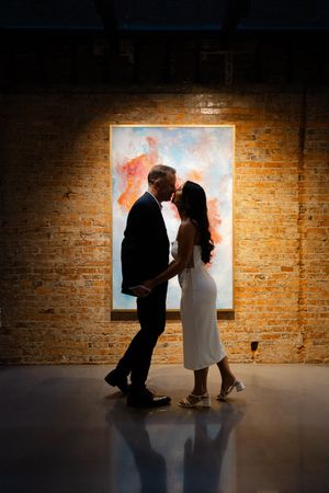 Silhouetted couple stands intimately in front of illuminated artwork on exposed brick wall in dark gallery setting.