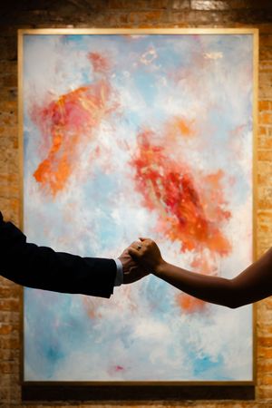 Two hands reach out to hold each other in front of an abstract painting with coral and blue tones.