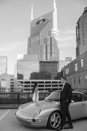 A couple stands next to a silver Porsche 911 in downtown Nashville with the AT&T Building in the background.