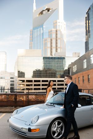 Silver Porsche sports car parked in urban setting with city skyline and parking garage in background.