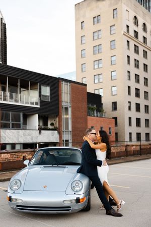 A couple embraces against a silver Porsche 911 parked in front of a modern urban building.