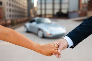 Two hands grasp in greeting with a silver luxury car parked in the blurred urban background.