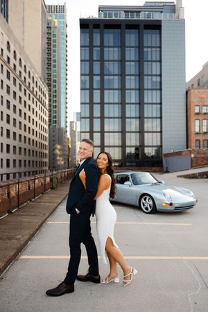 A couple poses romantically in front of modern urban architecture with a silver Porsche sports car in the background.