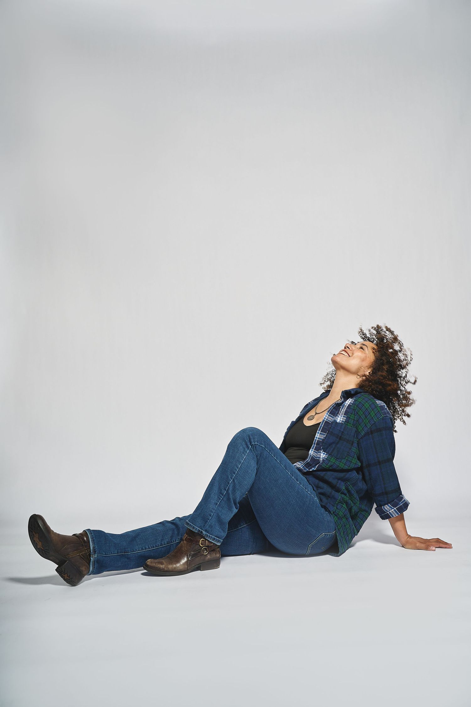 A woman in jeans and plaid shirt sitting on the floor with head tilted back on white background.