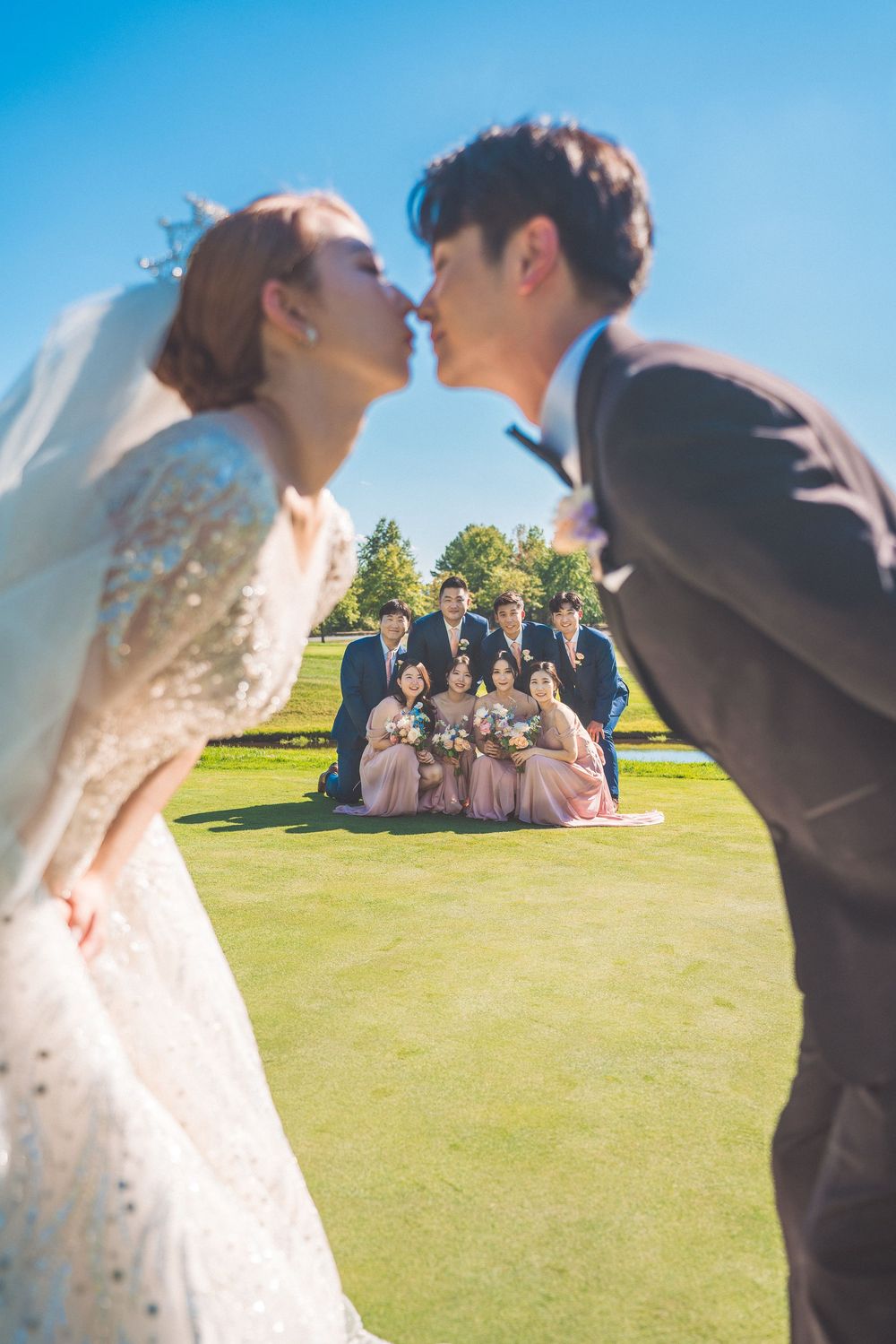 Creative wedding photo with couple in foreground and wedding party in background on grass.