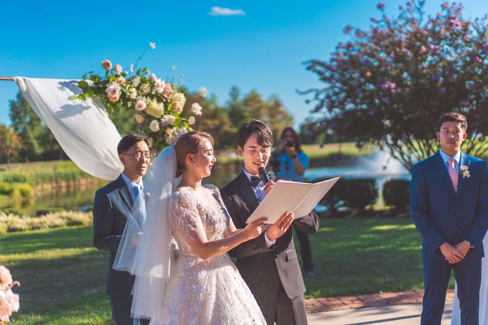 Outdoor wedding ceremony with couple and officiant reading from paper in sunlit garden.