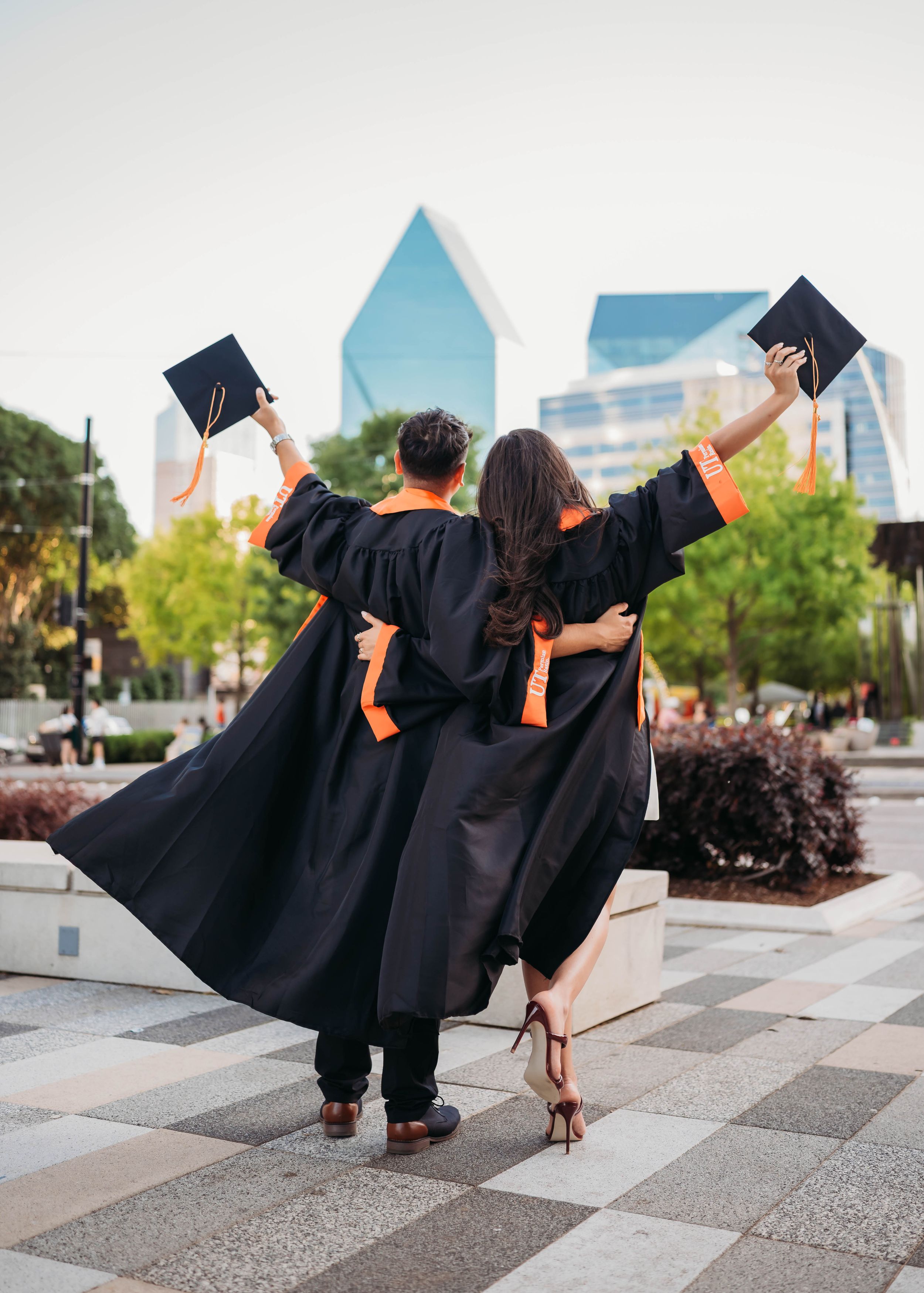 Two graduates in black caps and gowns raise their mortarboards while celebrating in front of modern city buildings.