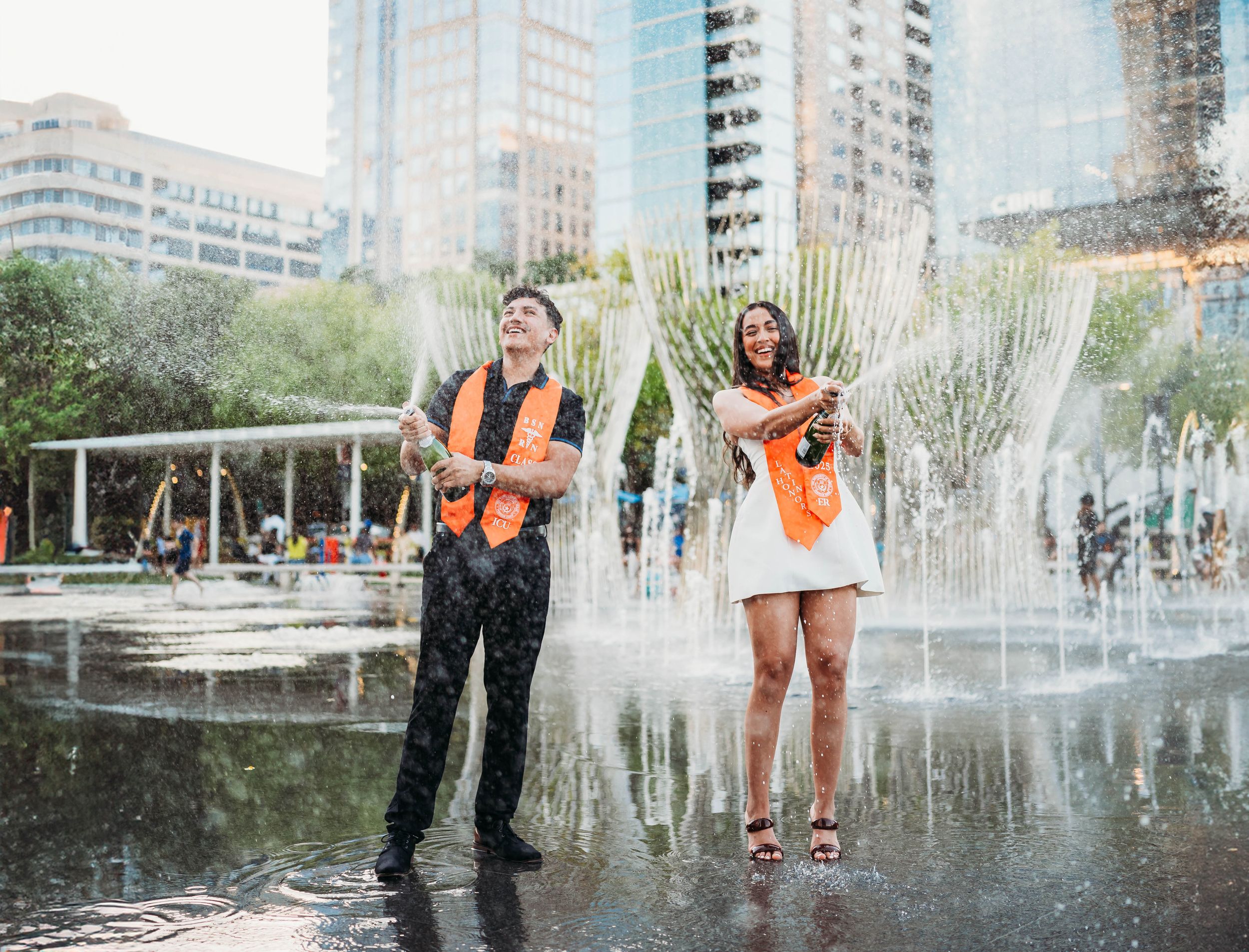 People in orange safety vests splash through shallow fountain water in front of modern city buildings.
