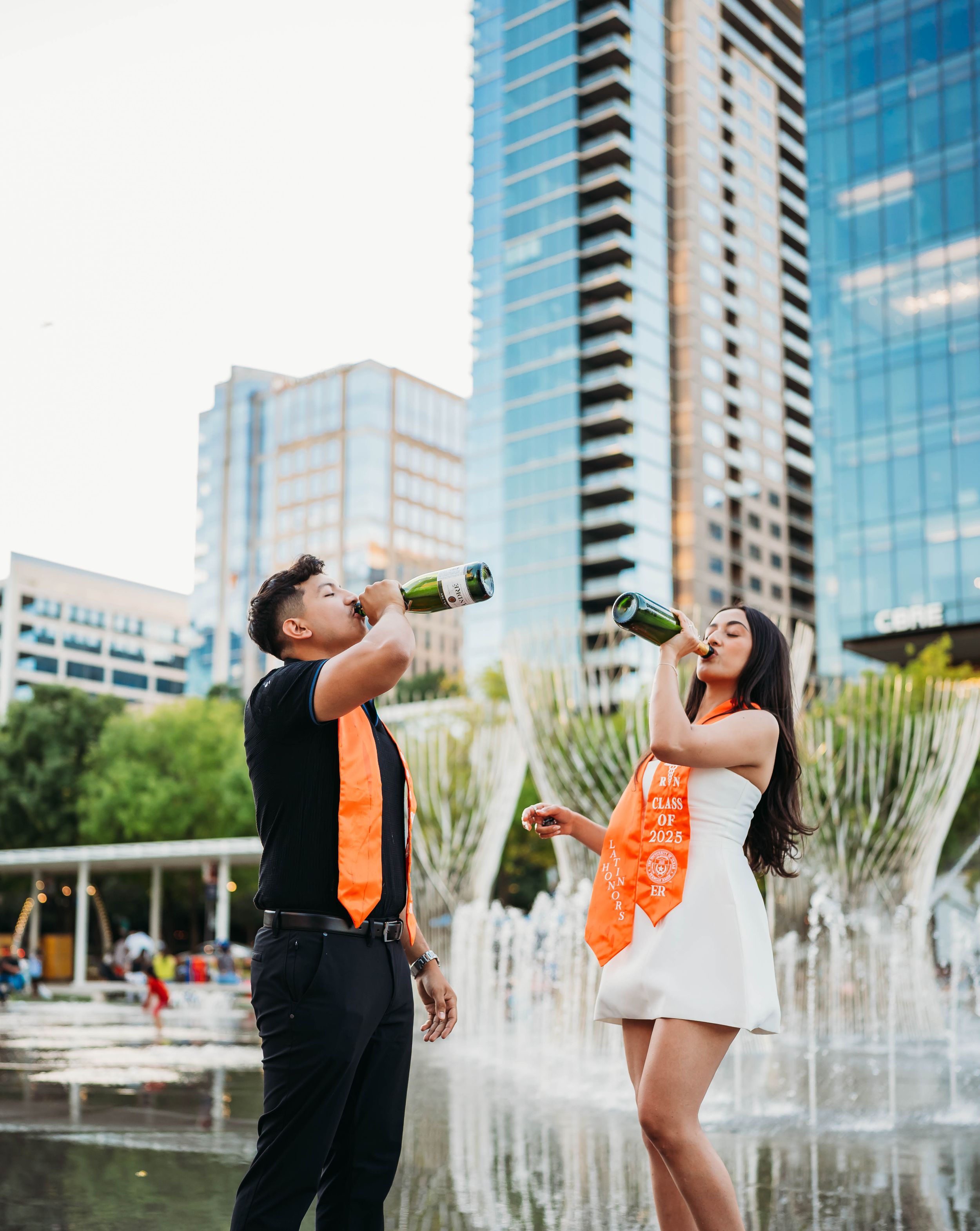 Couple pops a champagne bottle and celebrates near fountains in an urban courtyard with modern buildings.