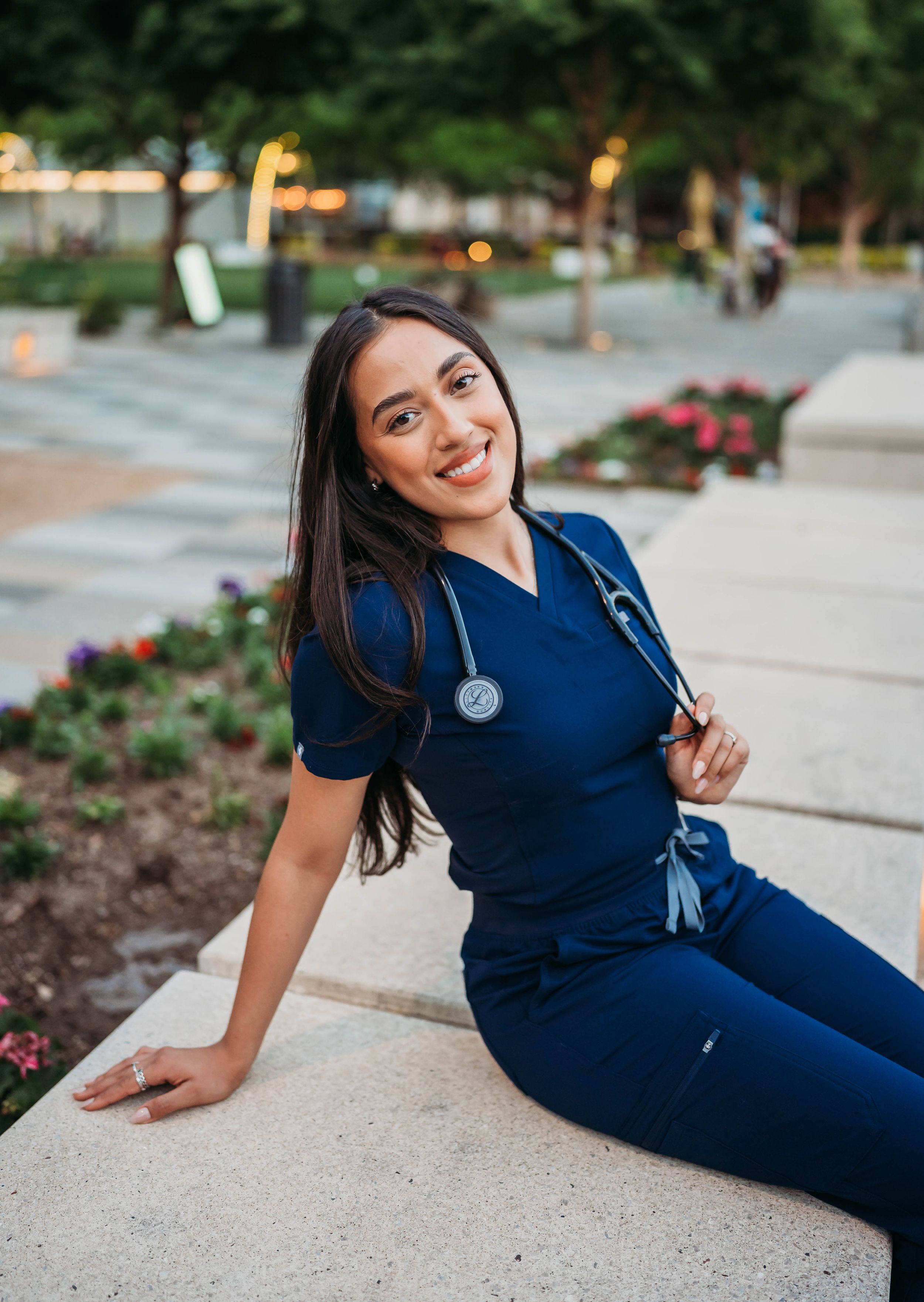 Healthcare professional in navy blue scrubs sitting outdoors near a floral garden during sunset.
