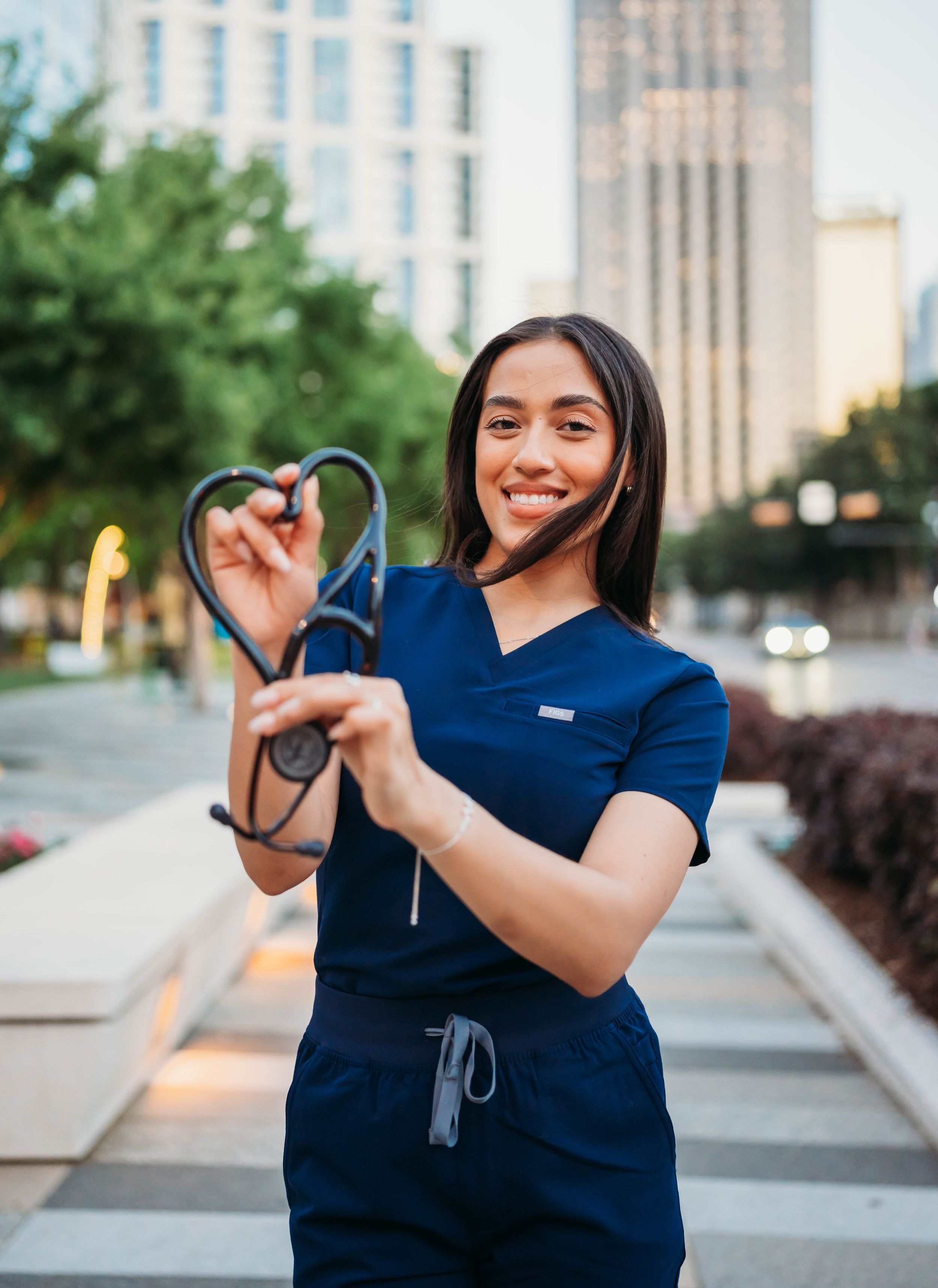 Healthcare professional in navy blue scrubs forms heart shape with stethoscope while smiling outdoors in urban setting.