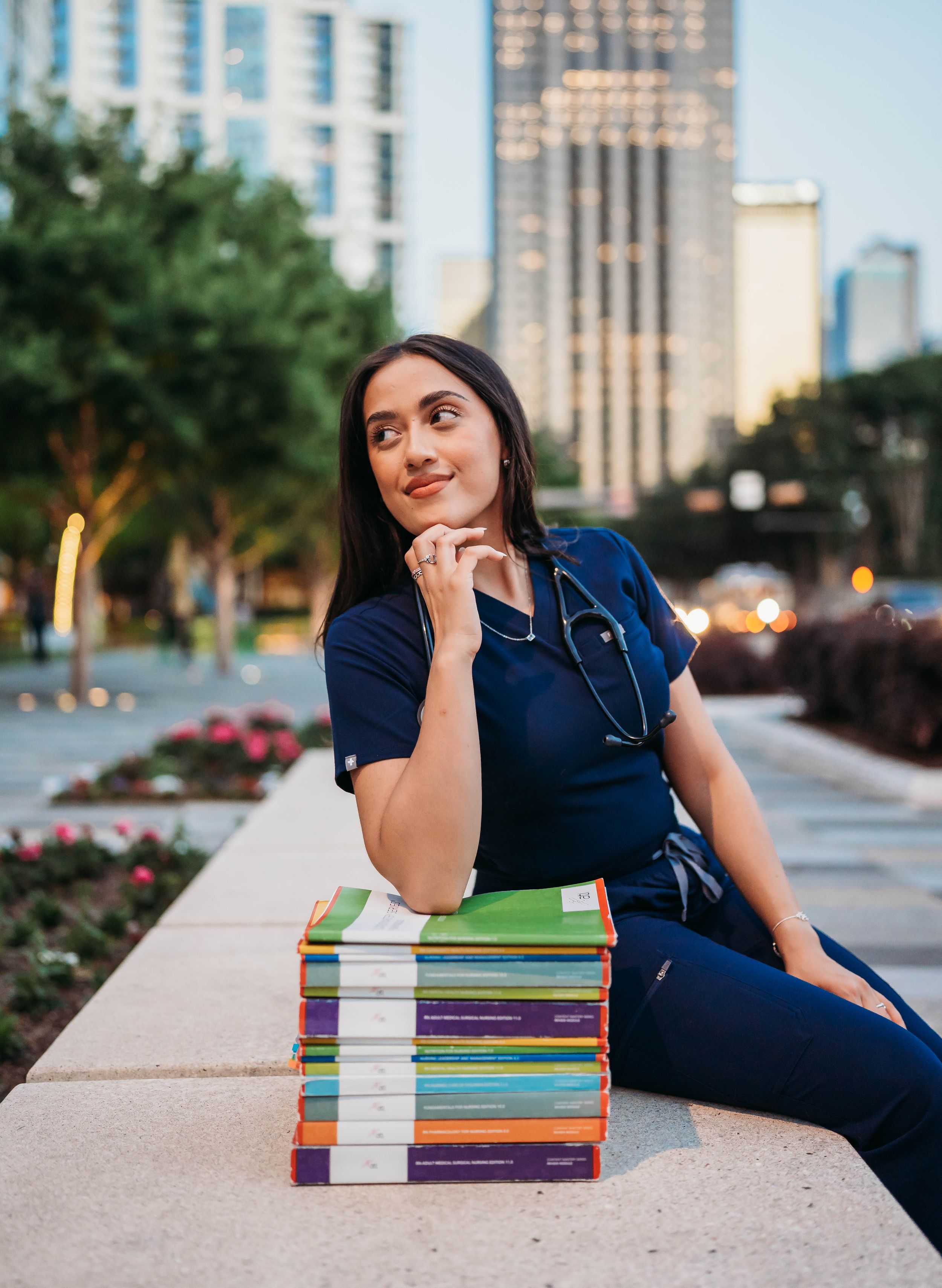 Medical student in navy blue scrubs rests thoughtfully on stack of colorful textbooks on urban campus ledge.
