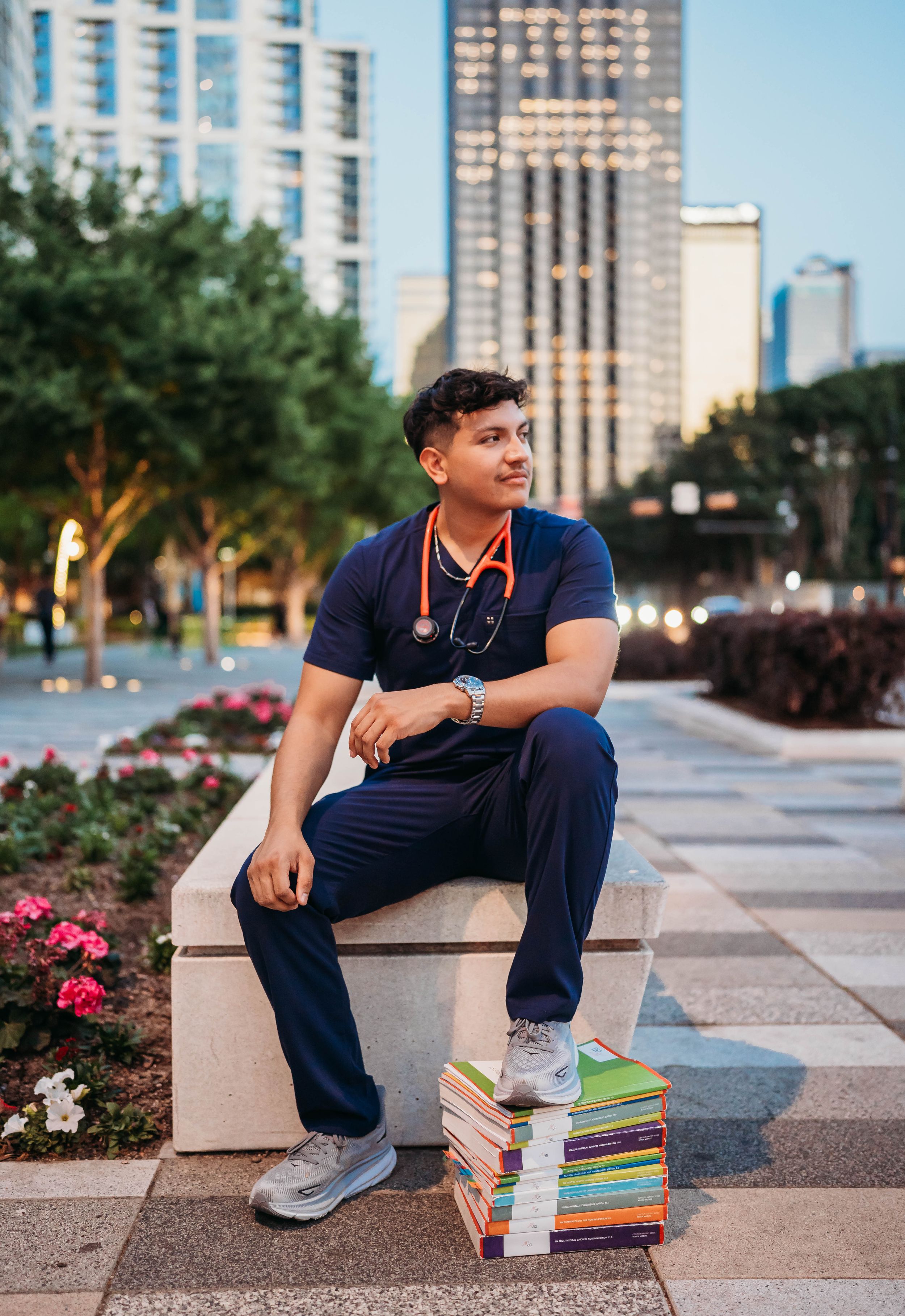 A person in navy scrubs sits contemplatively on stone steps with textbooks in an urban setting at dusk.
