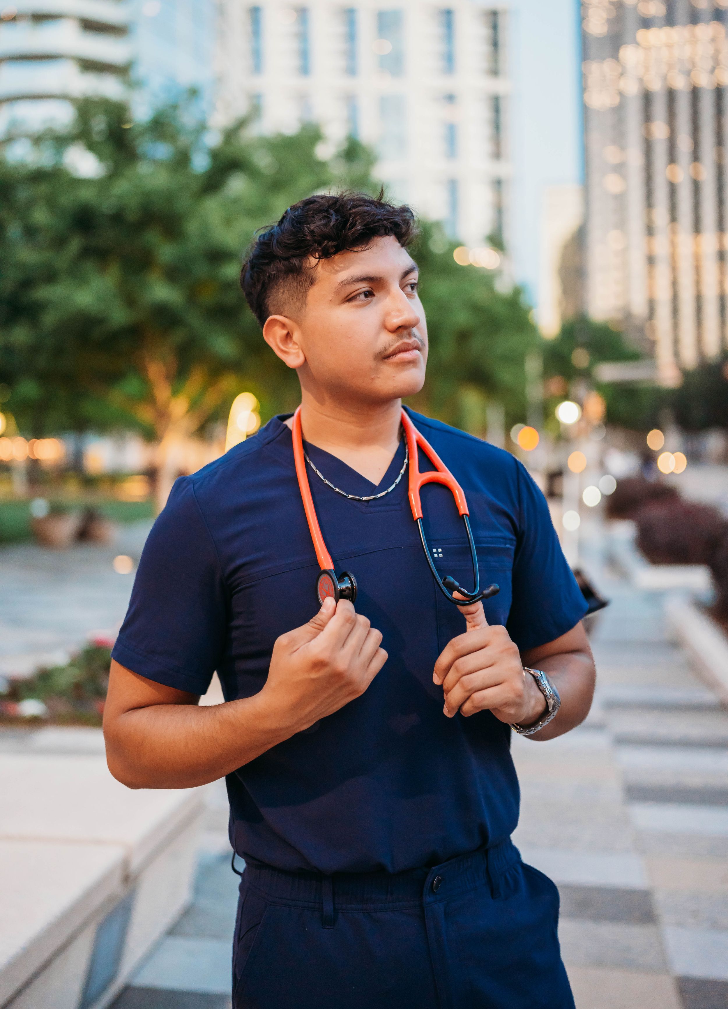 Healthcare worker in navy scrubs poses confidently with stethoscope against city backdrop in evening light.
