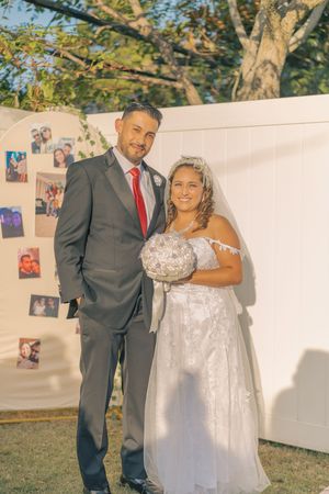 Wedding couple poses against white backdrop with personal photos displayed outdoors.