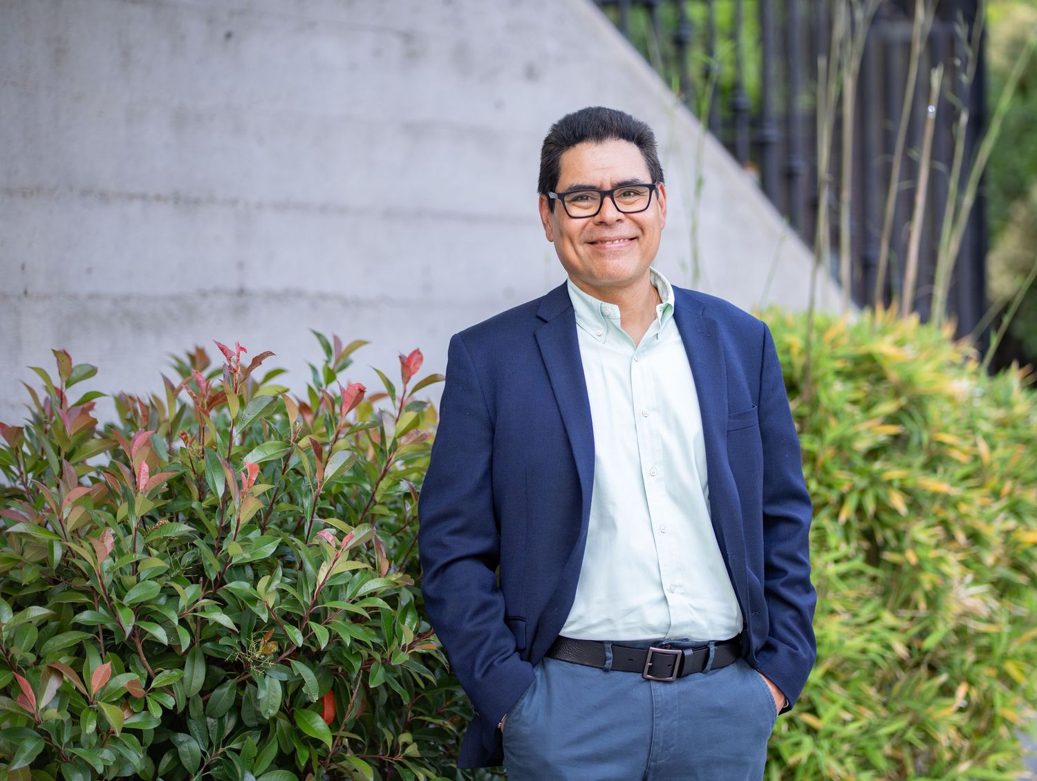 Professional wearing a navy blazer and glasses stands confidently by landscaped garden wall with hands in pockets.
