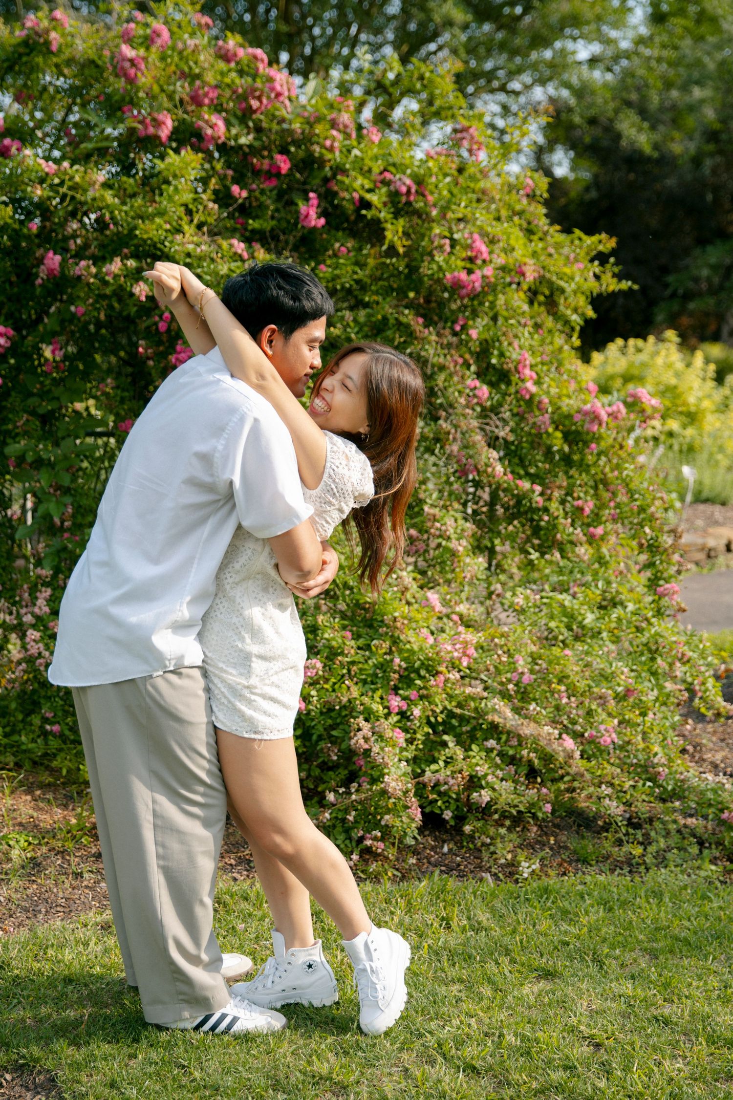 Two people in light colored outfits share an intimate moment against a backdrop of pink blooming roses in a garden setting.