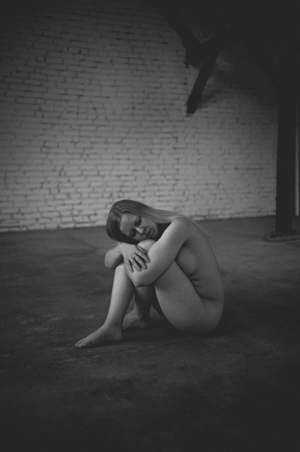 Artistic black and white nude figure curled up on dark floor against white brick wall in moody indoor setting.