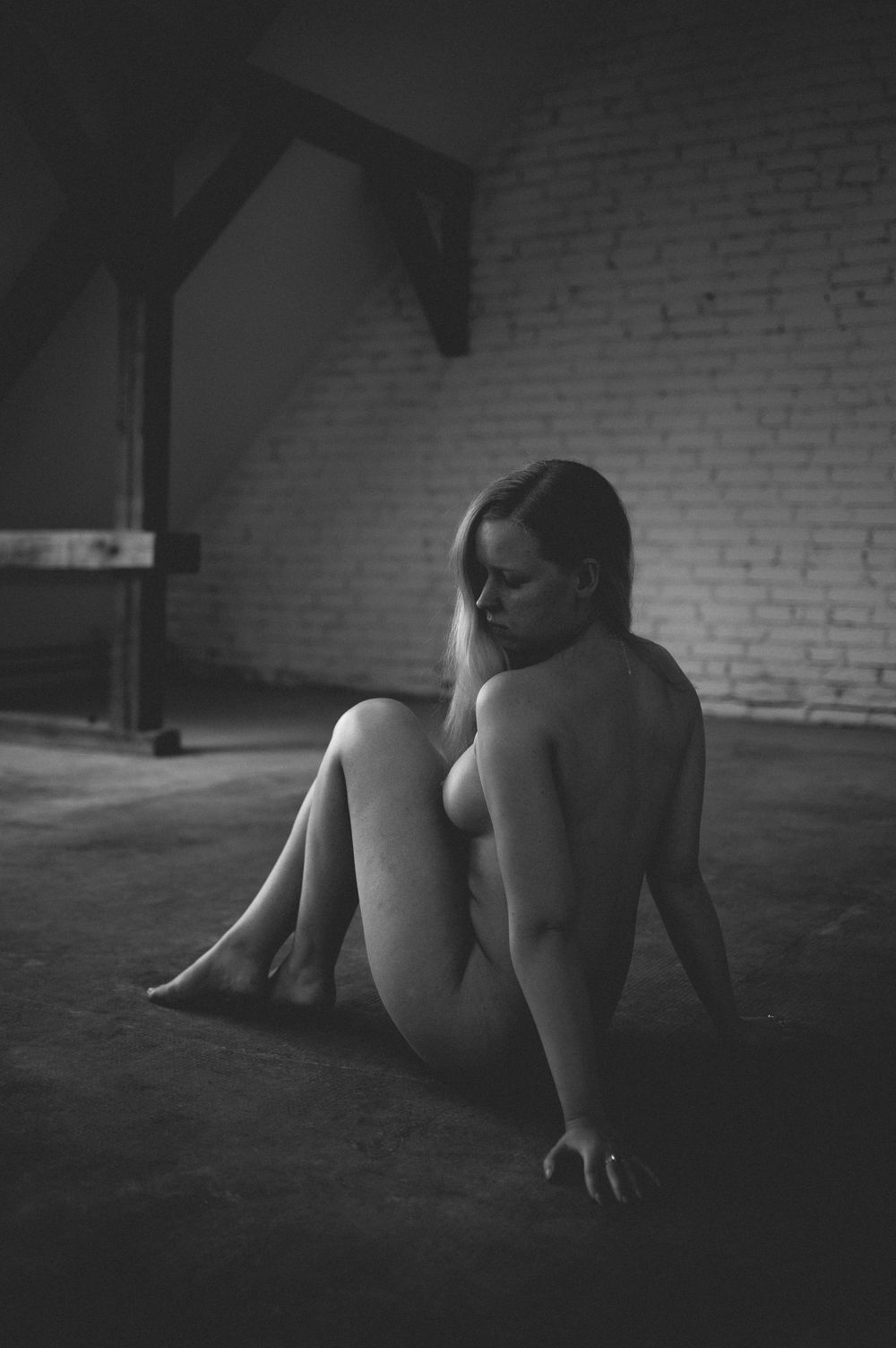 Black and white artistic nude photograph showing a figure sitting on dark floor against a brick wall with dramatic shadows.