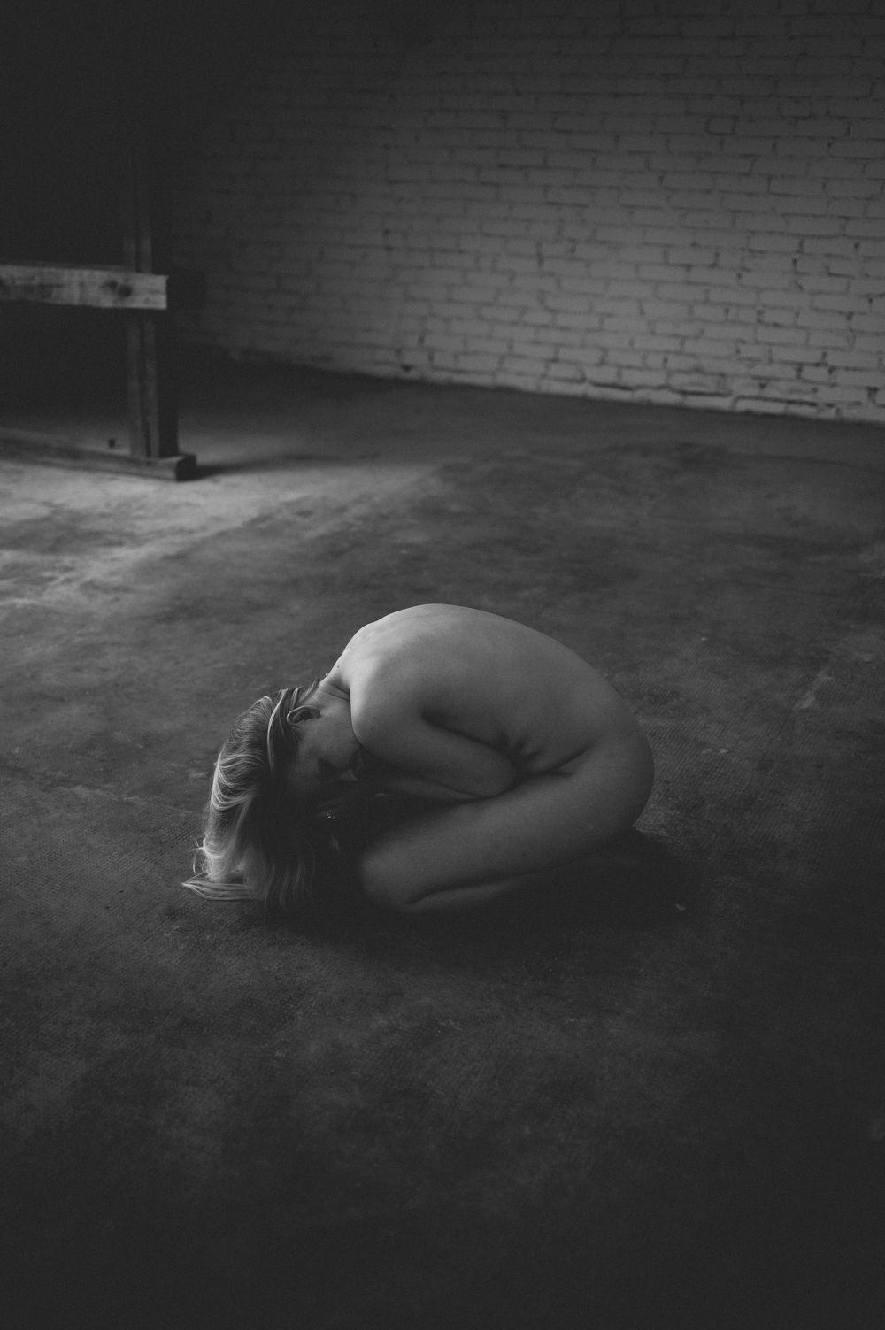 Black and white artistic photograph of a crouched figure in a bare industrial room with brick walls and concrete floor.
