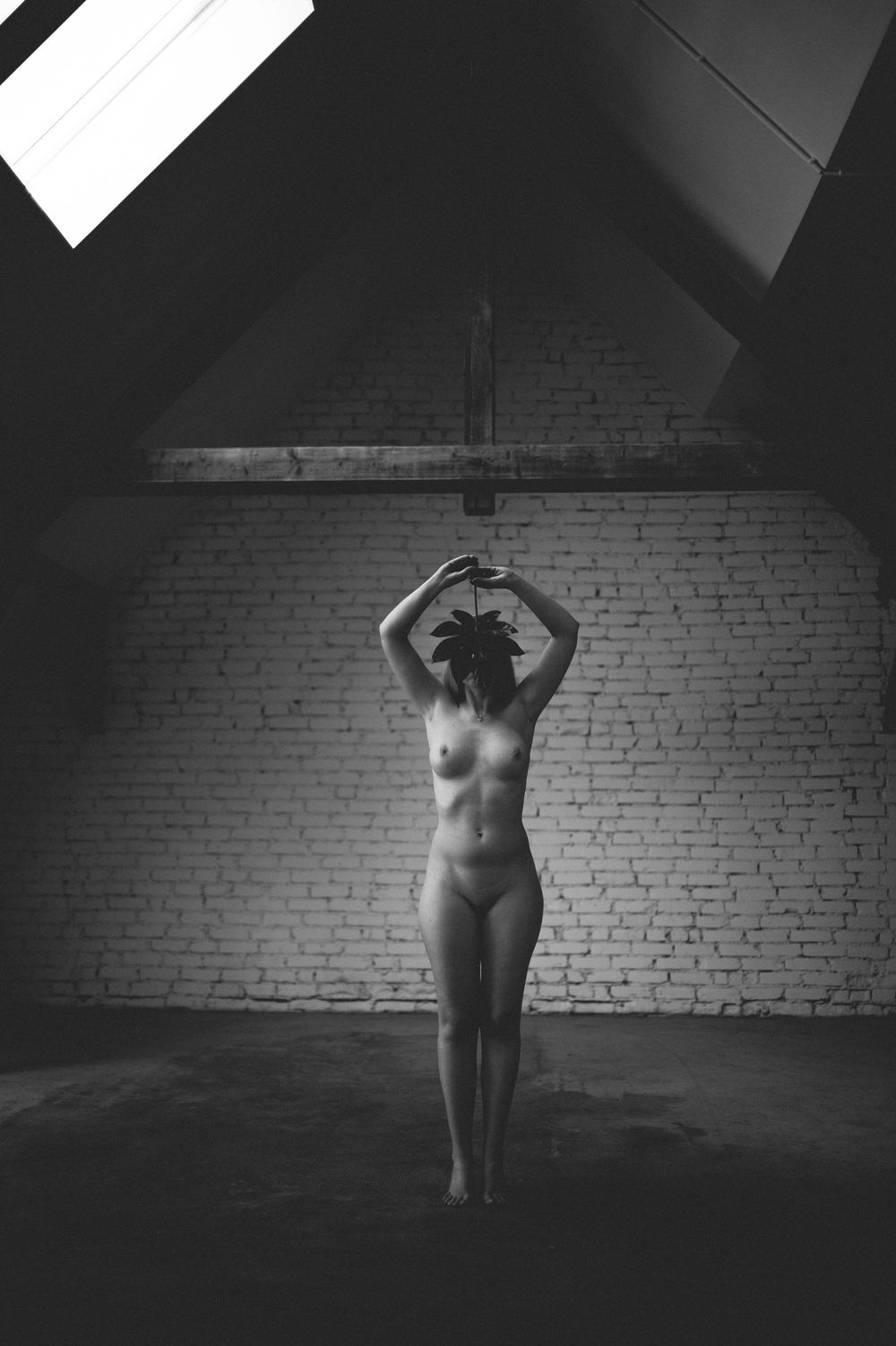 A black and white artistic nude photograph of a figure in silhouette against a white brick wall in an attic space.