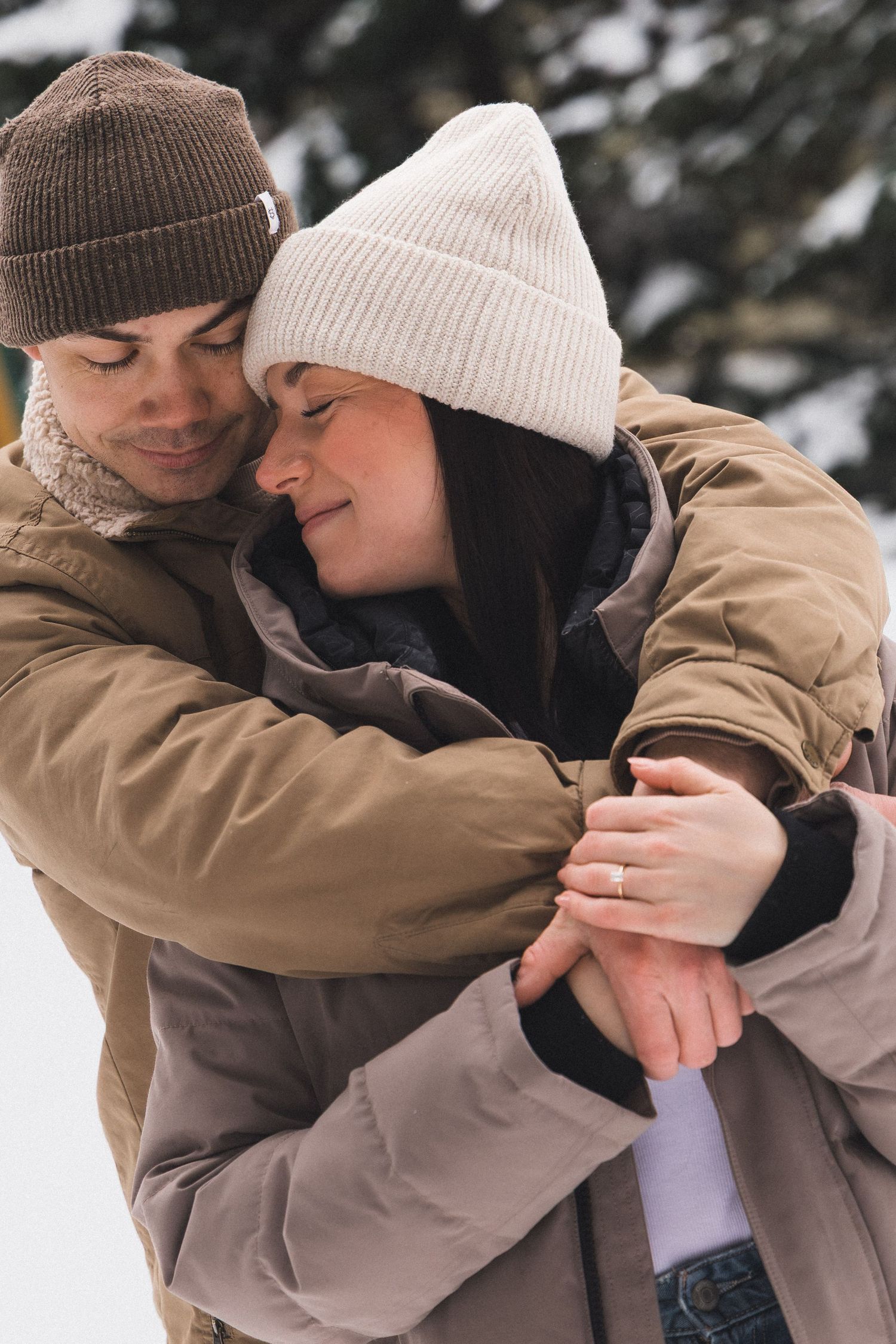 Happy couple embracing in snowy winter landscape wearing warm coats and beanies, woman displaying engagement ring.