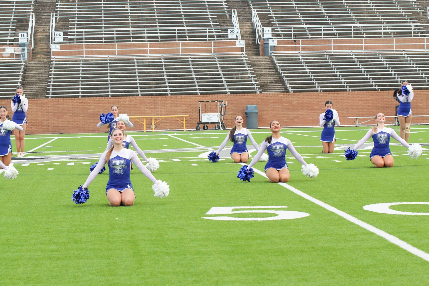 Football players stretch and warm up on a green turf field with empty bleachers in the background.