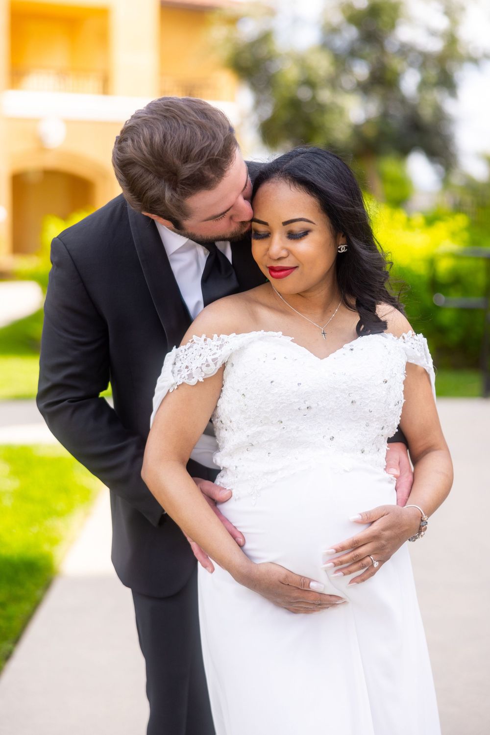 A romantic maternity photo showing a couple embracing outdoors, with the pregnant woman wearing an elegant white lace dress.