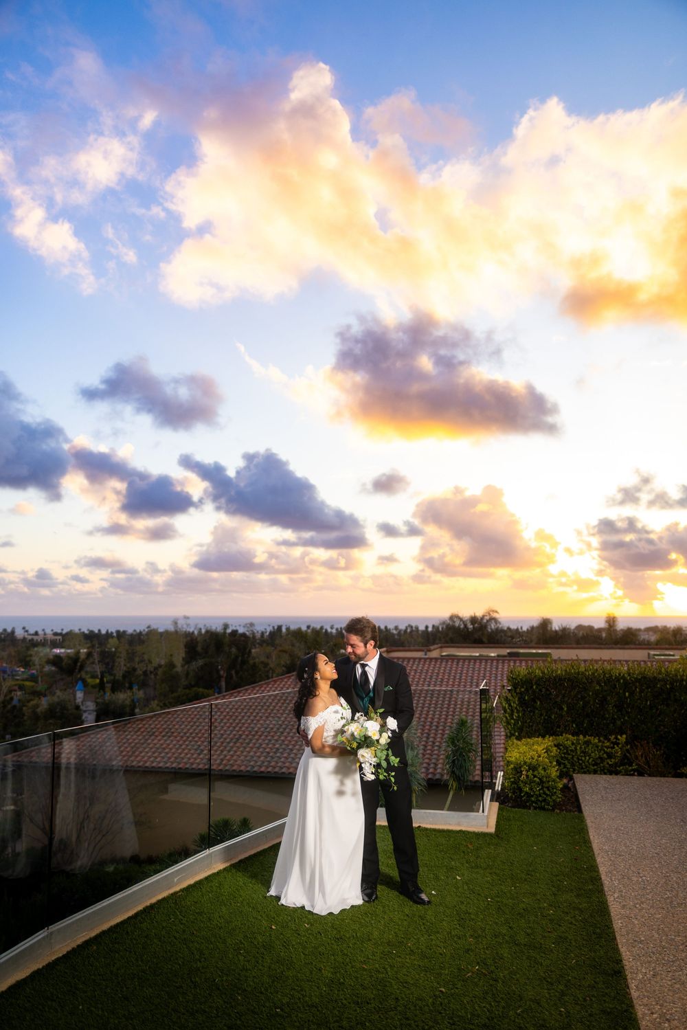 A couple shares a romantic moment on a rooftop terrace with a stunning sunset and cloudy sky in the background.