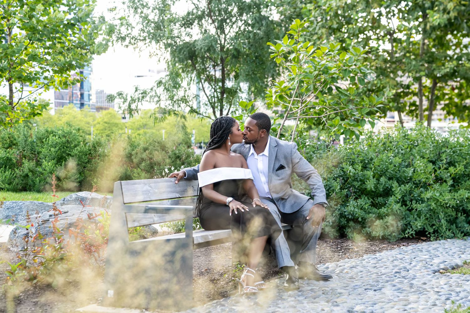 A couple sits together on a park bench surrounded by lush greenery and landscaping on a sunny afternoon.