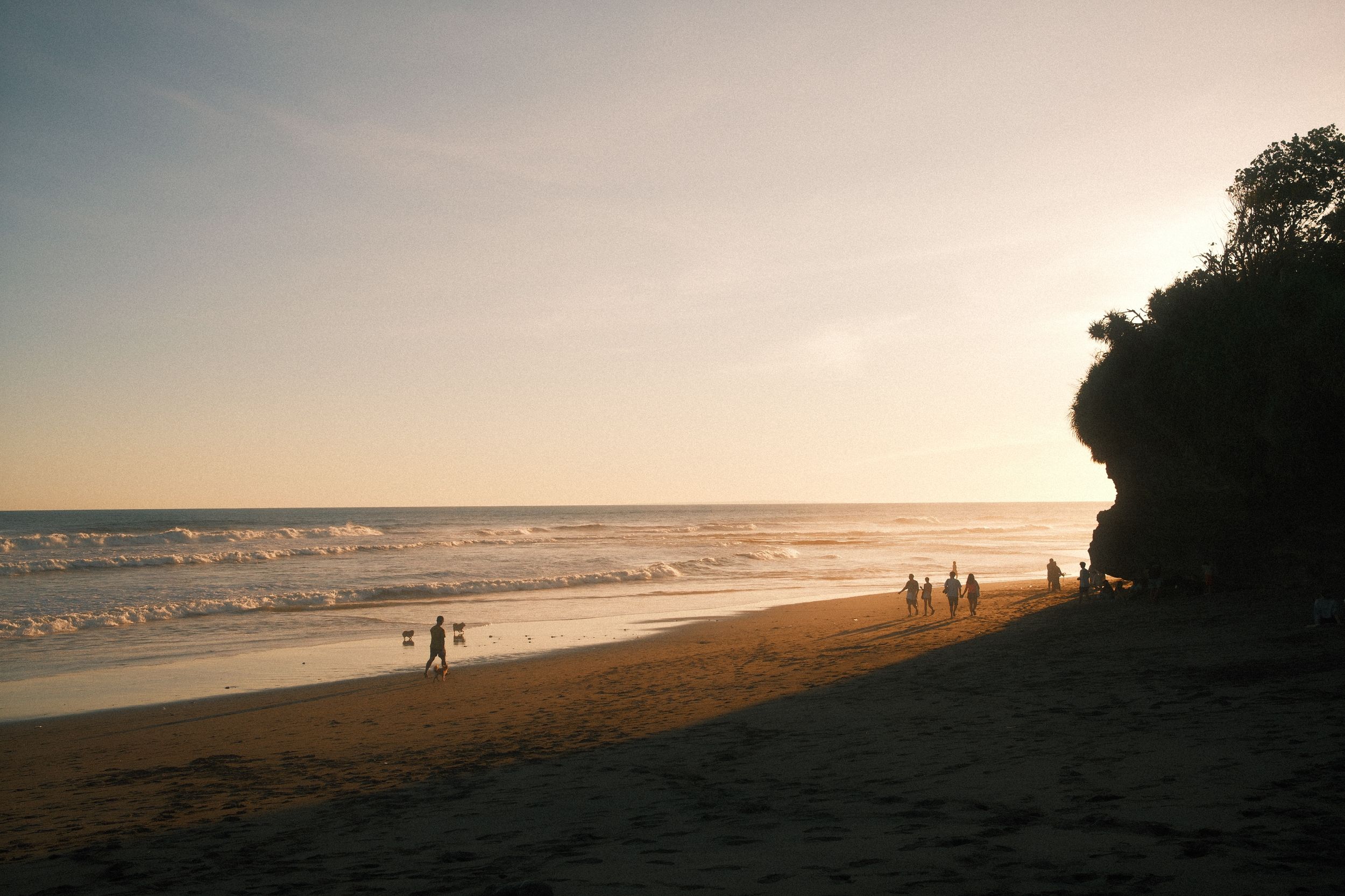 Silhouettes of people walking along a golden sunset beach with dramatic coastal cliffs in the background.