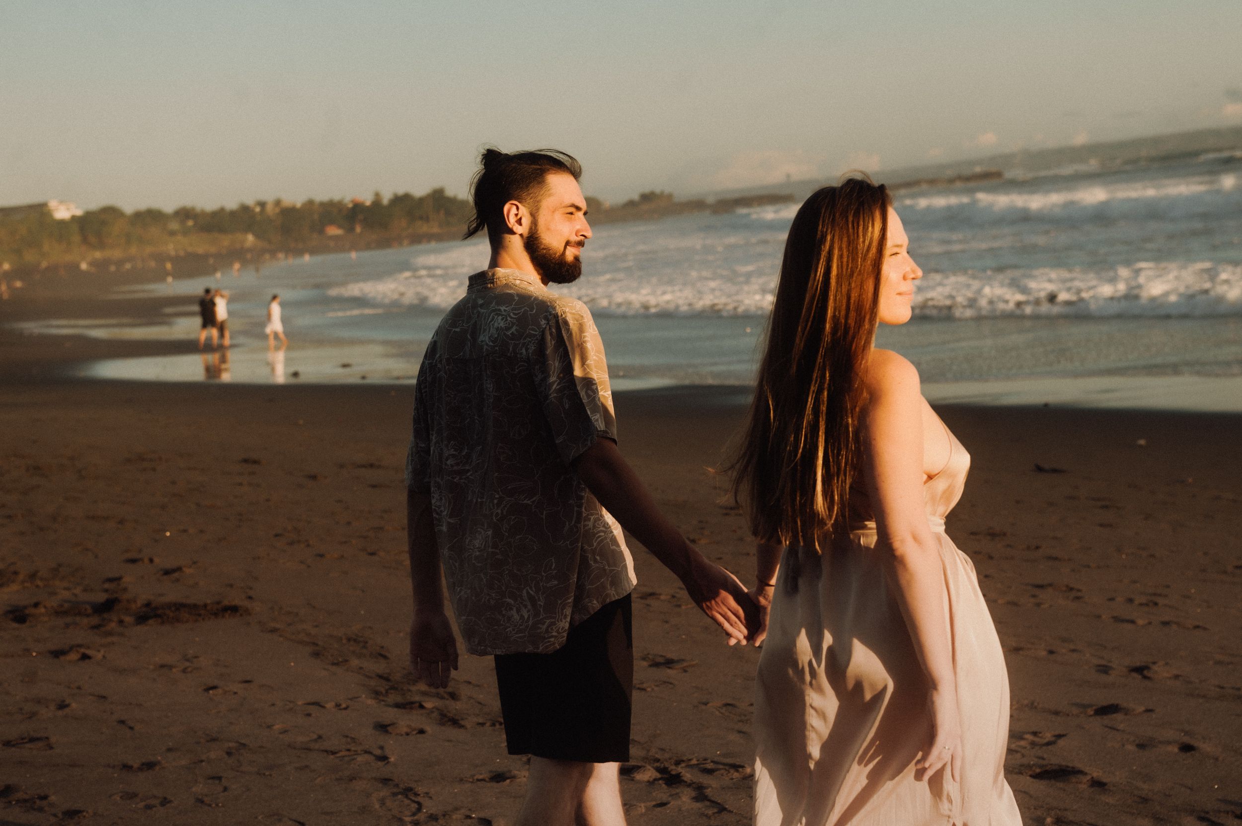 A couple walks along a sandy beach at sunset with waves crashing in the background.